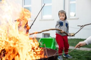 cute little adorable baby boy put big wooden stick in fire pit with huge fire at house garden yard