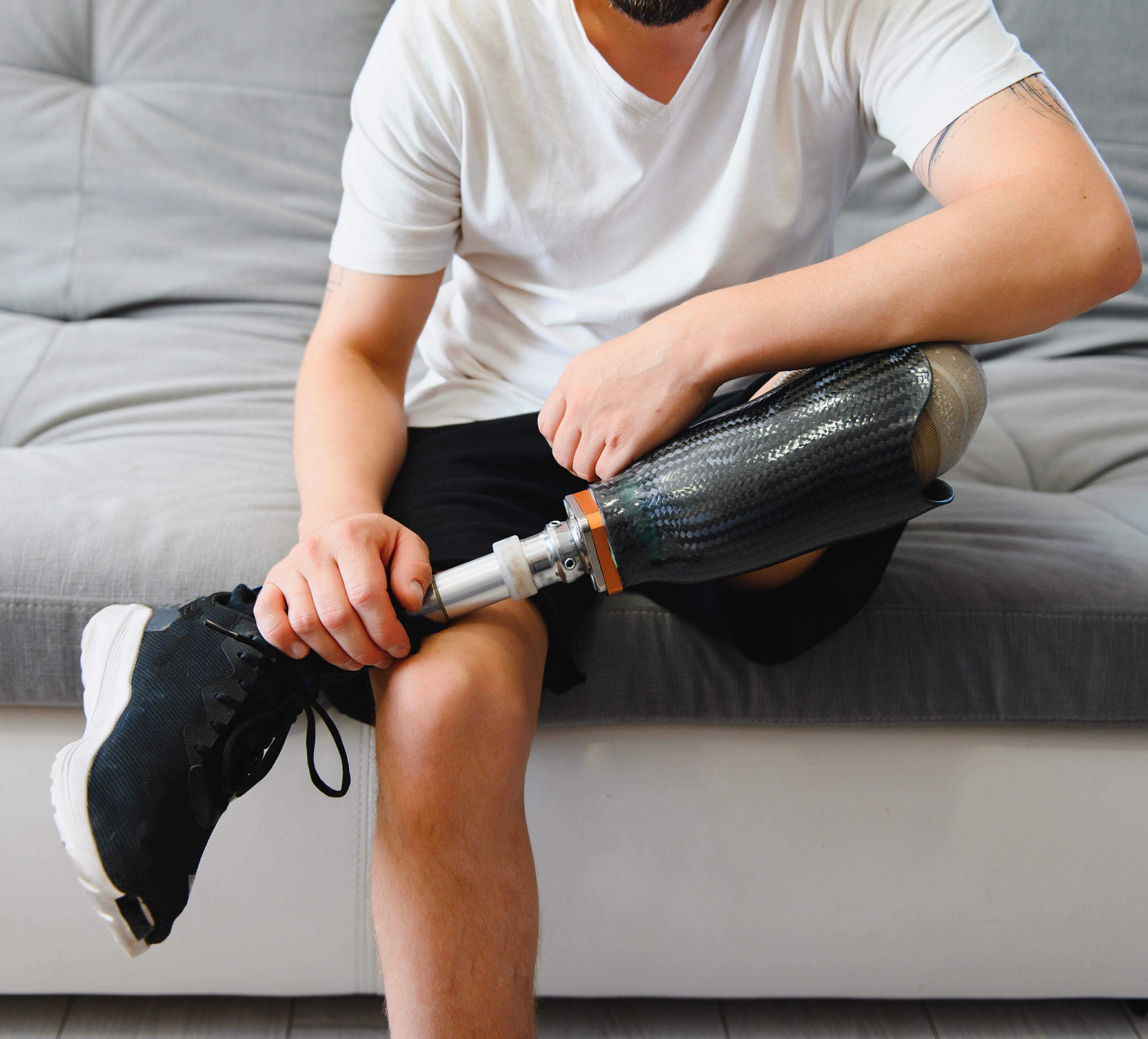 Man amputee with prosthetic leg disability on above knee transfemoral leg prosthesis wearing shoe sitting on sofa, close up. People with amputation disabilities in everyday life