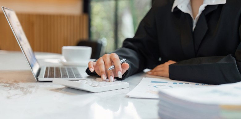 A woman with an open laptop and papers on her desk use a calculator to add up numbers